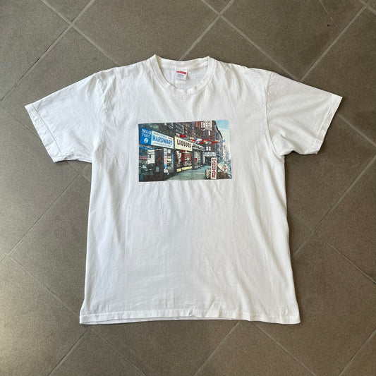 Supreme SS18 Hardware White Tee features a white 100% cotton T-shirt made in the USA. The front has a street scene with various storefronts, including a hardware store with a Supreme logo above it. A small Supreme box logo is printed on the upper back below the collar.

