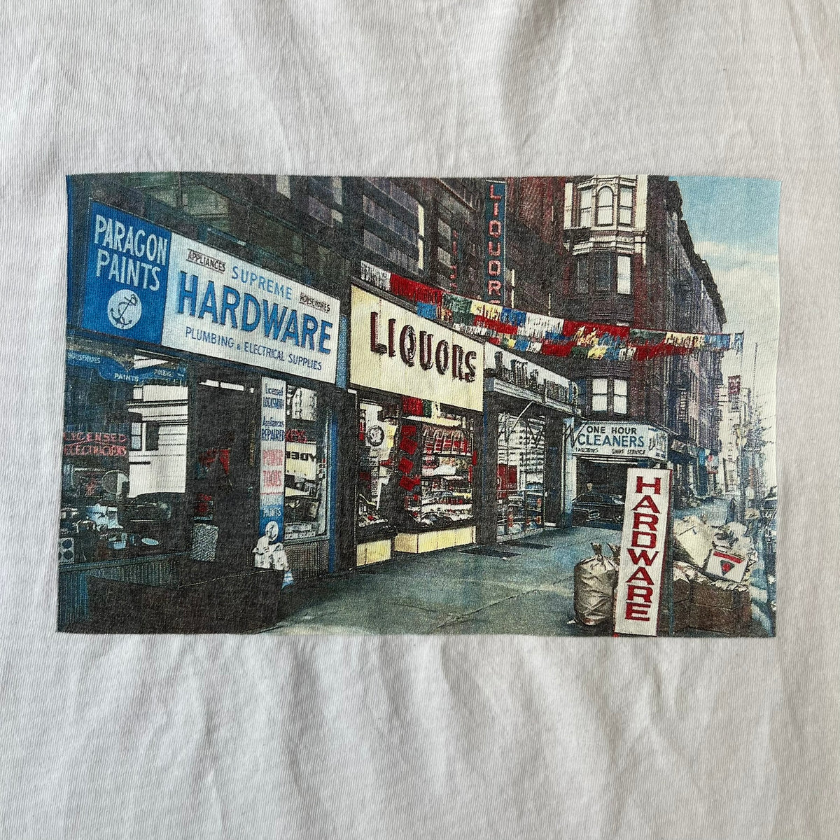 Supreme SS18 Hardware White Tee features a white 100% cotton T-shirt made in the USA. The front has a street scene with various storefronts, including a hardware store with a Supreme logo above it. A small Supreme box logo is printed on the upper back below the collar.

