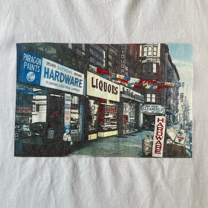 Supreme SS18 Hardware White Tee features a white 100% cotton T-shirt made in the USA. The front has a street scene with various storefronts, including a hardware store with a Supreme logo above it. A small Supreme box logo is printed on the upper back below the collar.
