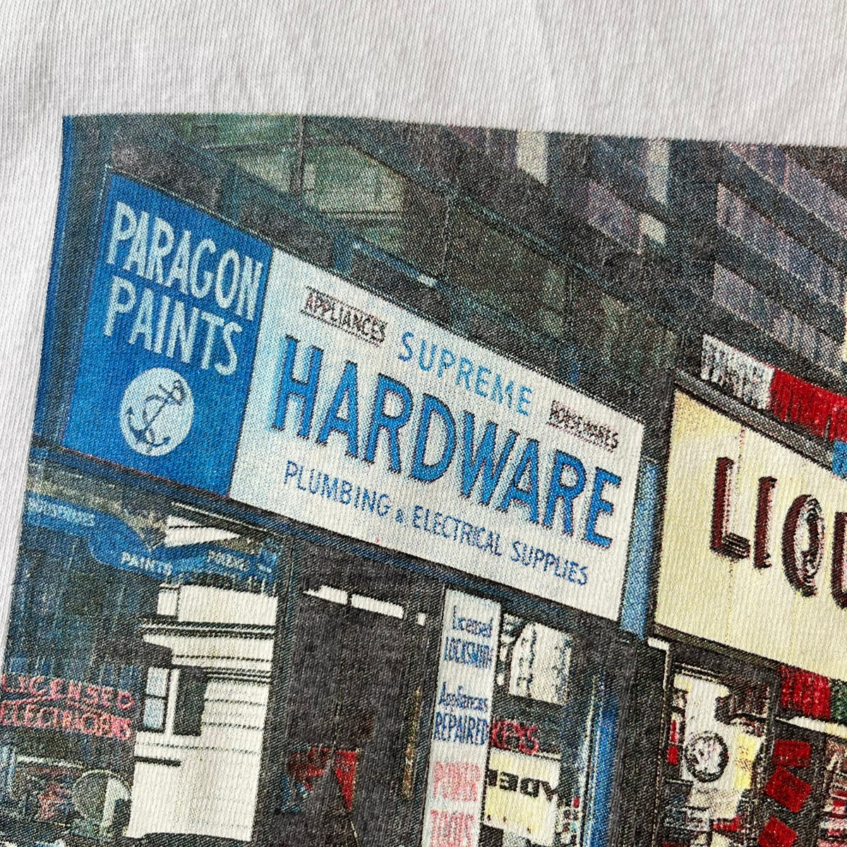 Supreme SS18 Hardware White Tee features a white 100% cotton T-shirt made in the USA. The front has a street scene with various storefronts, including a hardware store with a Supreme logo above it. A small Supreme box logo is printed on the upper back below the collar.
