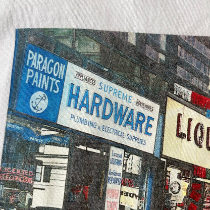 Supreme SS18 Hardware White Tee features a white 100% cotton T-shirt made in the USA. The front has a street scene with various storefronts, including a hardware store with a Supreme logo above it. A small Supreme box logo is printed on the upper back below the collar.
