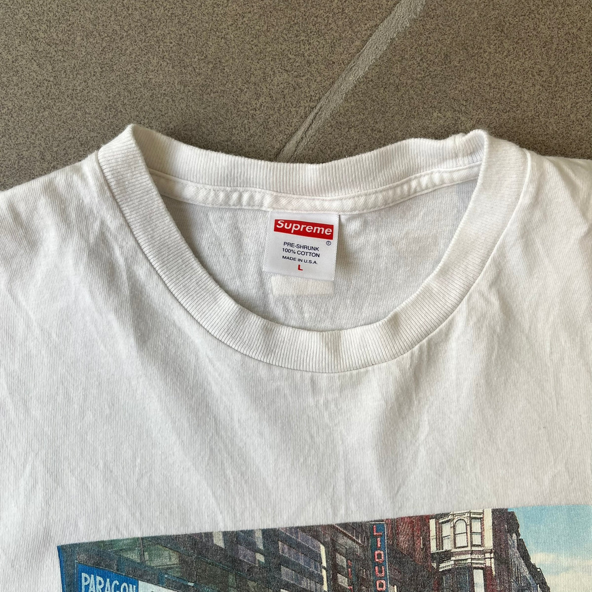 Supreme SS18 Hardware White Tee features a white 100% cotton T-shirt made in the USA. The front has a street scene with various storefronts, including a hardware store with a Supreme logo above it. A small Supreme box logo is printed on the upper back below the collar.
