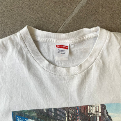 Supreme SS18 Hardware White Tee features a white 100% cotton T-shirt made in the USA. The front has a street scene with various storefronts, including a hardware store with a Supreme logo above it. A small Supreme box logo is printed on the upper back below the collar.
