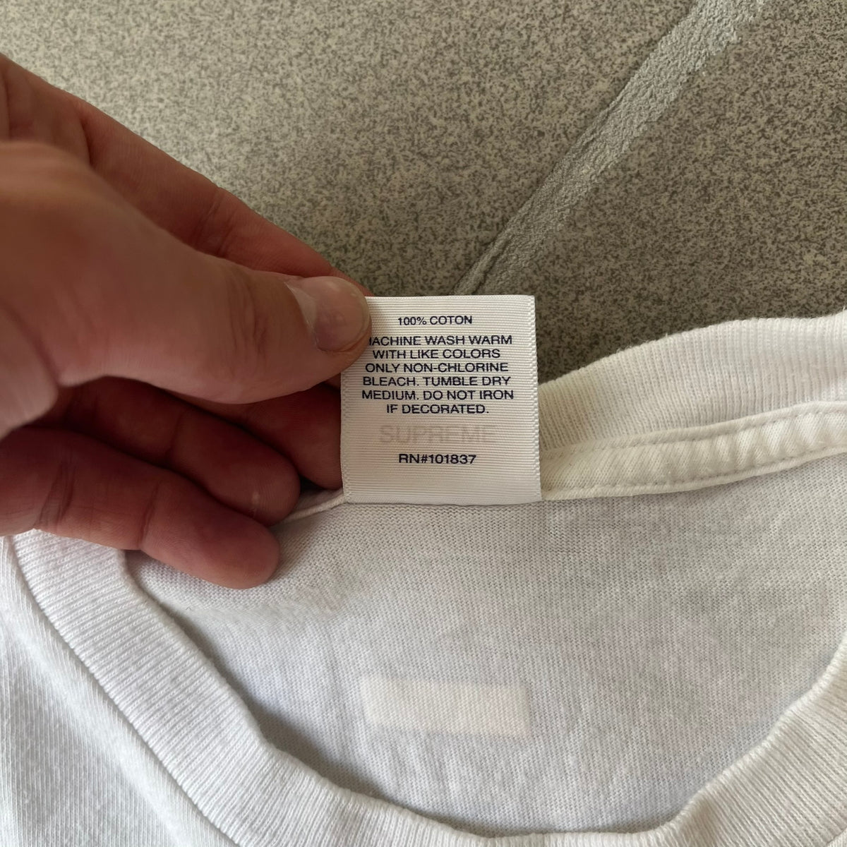 Supreme SS18 Hardware White Tee features a white 100% cotton T-shirt made in the USA. The front has a street scene with various storefronts, including a hardware store with a Supreme logo above it. A small Supreme box logo is printed on the upper back below the collar.
