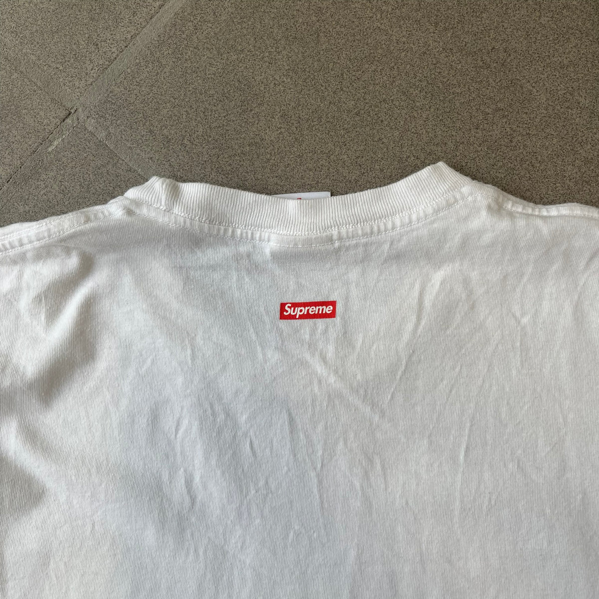 Supreme SS18 Hardware White Tee features a white 100% cotton T-shirt made in the USA. The front has a street scene with various storefronts, including a hardware store with a Supreme logo above it. A small Supreme box logo is printed on the upper back below the collar.
