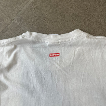 Supreme SS18 Hardware White Tee features a white 100% cotton T-shirt made in the USA. The front has a street scene with various storefronts, including a hardware store with a Supreme logo above it. A small Supreme box logo is printed on the upper back below the collar.

