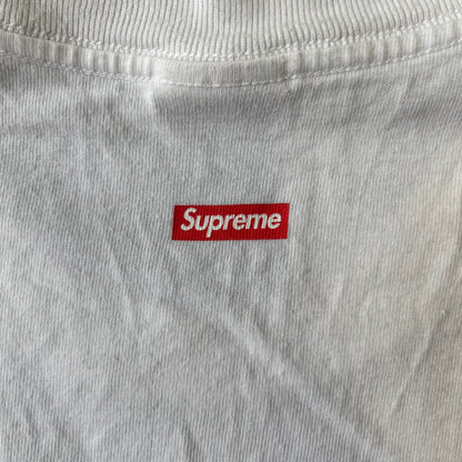 Supreme SS18 Hardware White Tee features a white 100% cotton T-shirt made in the USA. The front has a street scene with various storefronts, including a hardware store with a Supreme logo above it. A small Supreme box logo is printed on the upper back below the collar.
