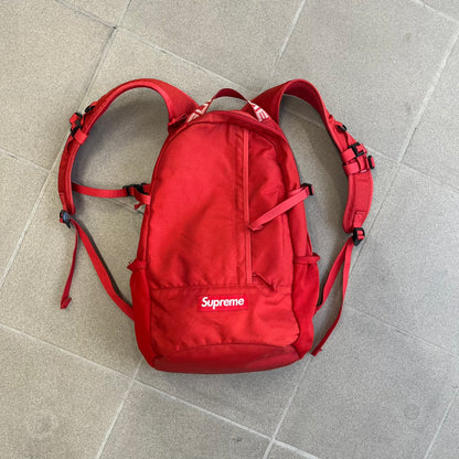 Supreme SS18 Red Backpack features a red construction with a large main compartment suitable for laptops and other larger devices, along with a front zippered pocket for smaller items. It has dual side mesh pockets and a Supreme Box Logo on the front.
