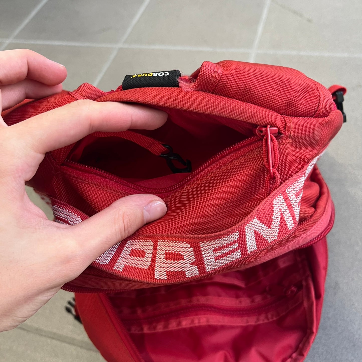 Supreme SS18 Red Backpack features a red construction with a large main compartment suitable for laptops and other larger devices, along with a front zippered pocket for smaller items. It has dual side mesh pockets and a Supreme Box Logo on the front.
