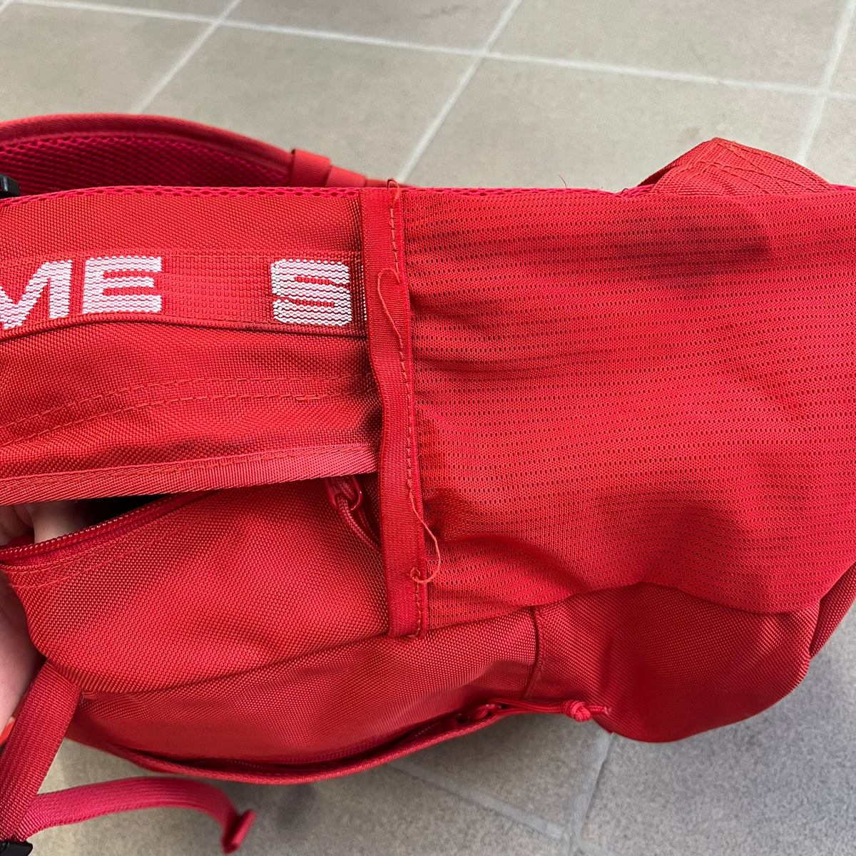 Supreme SS18 Red Backpack features a red construction with a large main compartment suitable for laptops and other larger devices, along with a front zippered pocket for smaller items. It has dual side mesh pockets and a Supreme Box Logo on the front.
