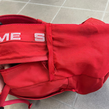 Supreme SS18 Red Backpack features a red construction with a large main compartment suitable for laptops and other larger devices, along with a front zippered pocket for smaller items. It has dual side mesh pockets and a Supreme Box Logo on the front.
