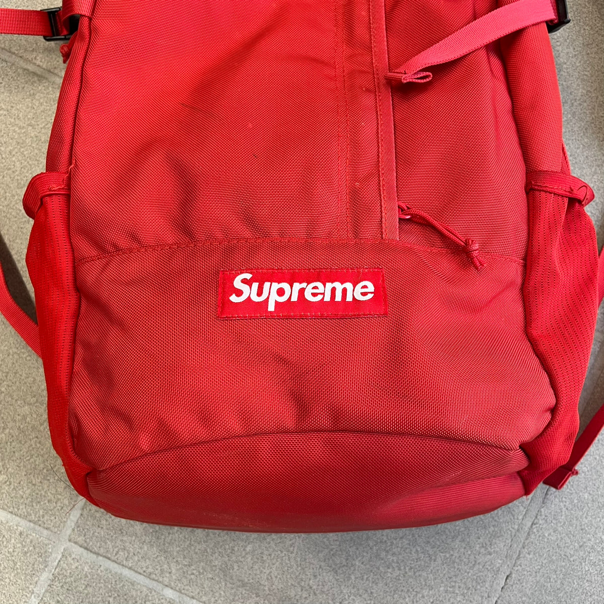 Supreme SS18 Red Backpack features a red construction with a large main compartment suitable for laptops and other larger devices, along with a front zippered pocket for smaller items. It has dual side mesh pockets and a Supreme Box Logo on the front.
