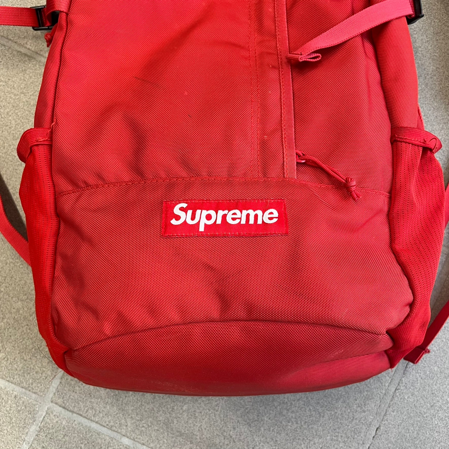 Supreme SS18 Red Backpack features a red construction with a large main compartment suitable for laptops and other larger devices, along with a front zippered pocket for smaller items. It has dual side mesh pockets and a Supreme Box Logo on the front.
