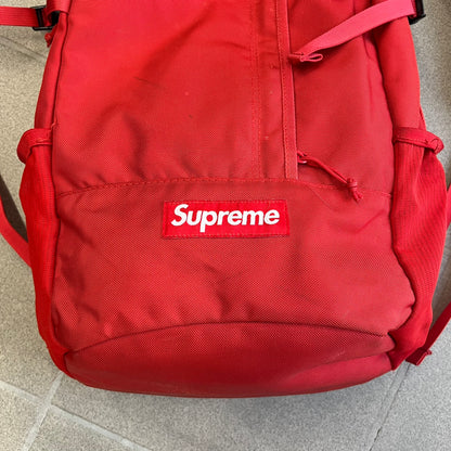 Supreme SS18 Red Backpack features a red construction with a large main compartment suitable for laptops and other larger devices, along with a front zippered pocket for smaller items. It has dual side mesh pockets and a Supreme Box Logo on the front.
