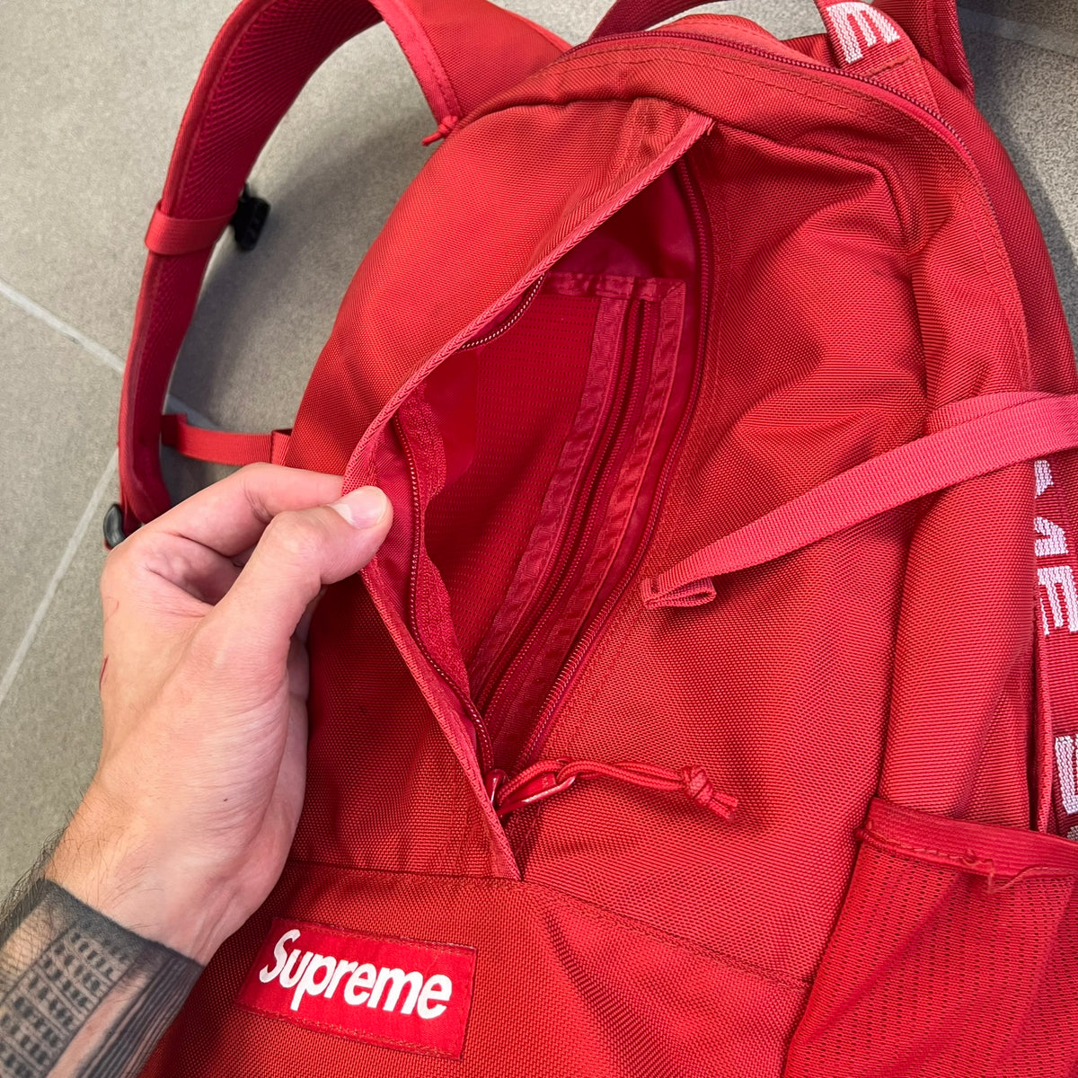 Supreme SS18 Red Backpack features a red construction with a large main compartment suitable for laptops and other larger devices, along with a front zippered pocket for smaller items. It has dual side mesh pockets and a Supreme Box Logo on the front.
