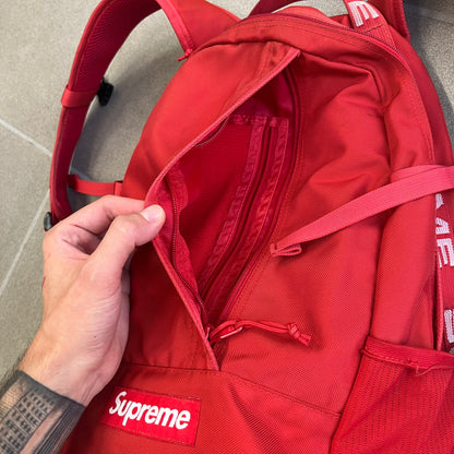 Supreme SS18 Red Backpack features a red construction with a large main compartment suitable for laptops and other larger devices, along with a front zippered pocket for smaller items. It has dual side mesh pockets and a Supreme Box Logo on the front.
