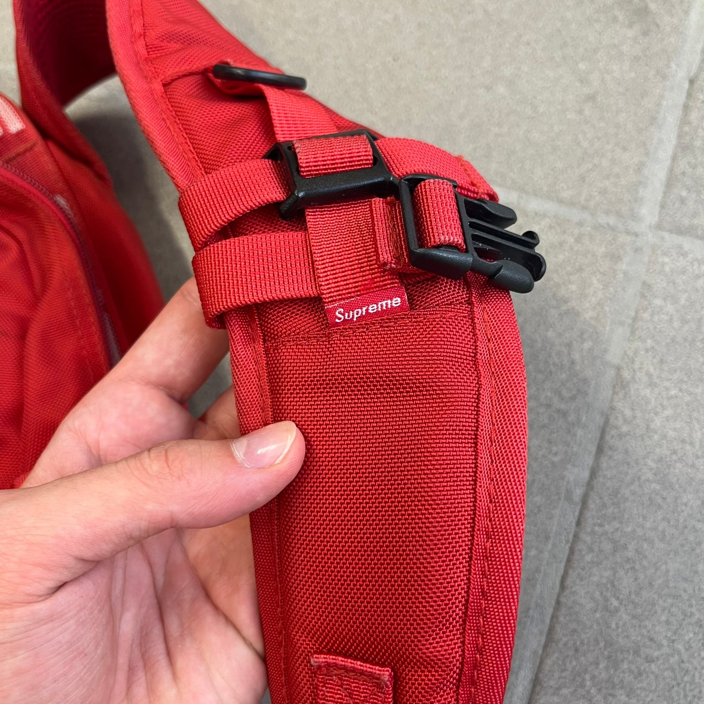 Supreme SS18 Red Backpack features a red construction with a large main compartment suitable for laptops and other larger devices, along with a front zippered pocket for smaller items. It has dual side mesh pockets and a Supreme Box Logo on the front.
