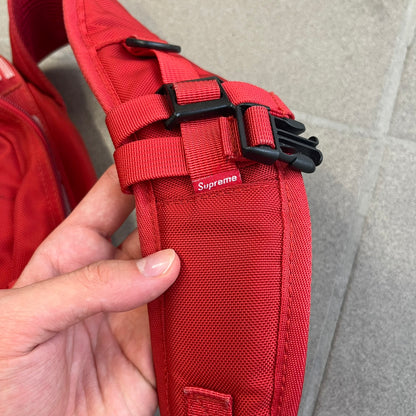 Supreme SS18 Red Backpack features a red construction with a large main compartment suitable for laptops and other larger devices, along with a front zippered pocket for smaller items. It has dual side mesh pockets and a Supreme Box Logo on the front.
