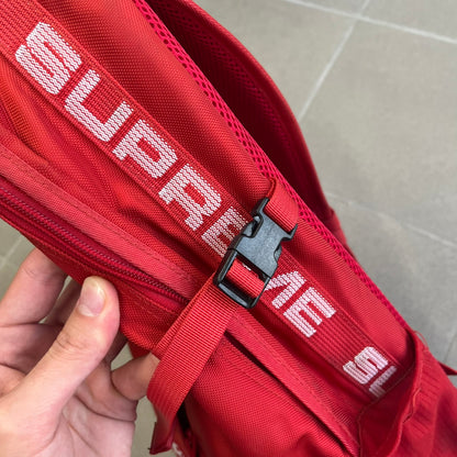 Supreme SS18 Red Backpack features a red construction with a large main compartment suitable for laptops and other larger devices, along with a front zippered pocket for smaller items. It has dual side mesh pockets and a Supreme Box Logo on the front.
