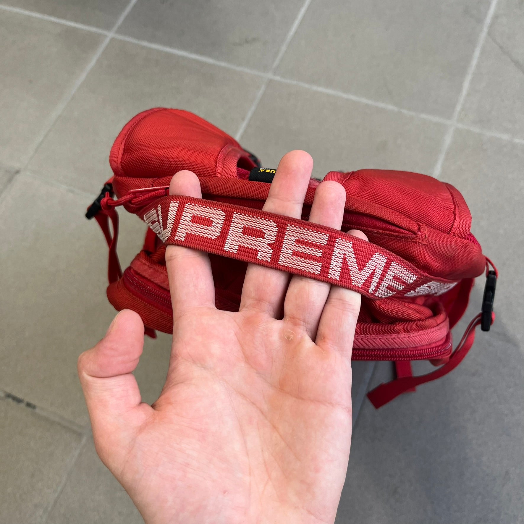 Supreme SS18 Red Backpack features a red construction with a large main compartment suitable for laptops and other larger devices, along with a front zippered pocket for smaller items. It has dual side mesh pockets and a Supreme Box Logo on the front.
