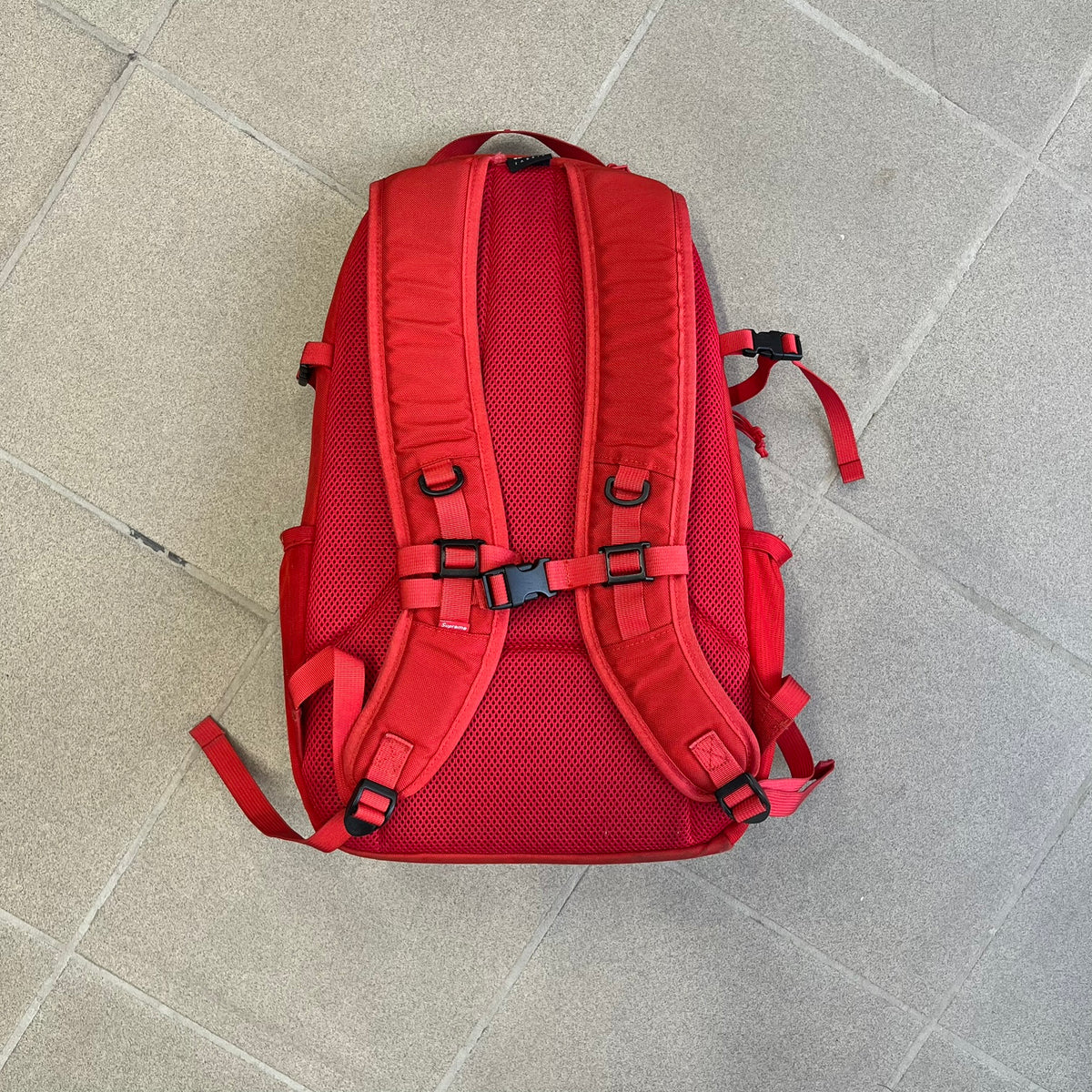 Supreme SS18 Red Backpack features a red construction with a large main compartment suitable for laptops and other larger devices, along with a front zippered pocket for smaller items. It has dual side mesh pockets and a Supreme Box Logo on the front.
