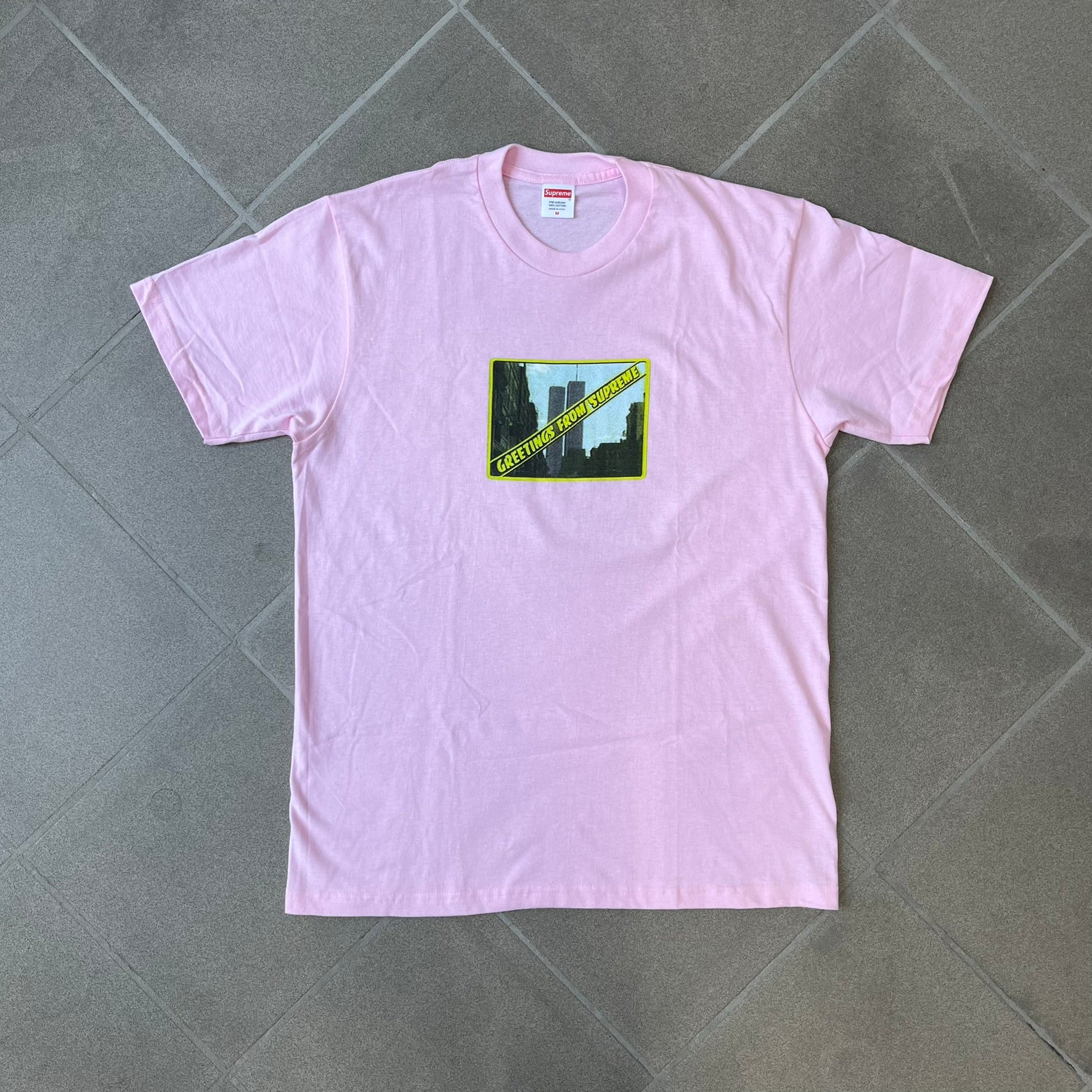 Supreme SS19 Greetings Pink Tee features a pink T-shirt with a front graphic designed as a greeting card showing the New York City skyline. The graphic includes the text “Greetings From Supreme” and depicts the original skyline with the Twin Towers. Released as part of Supreme’s Spring/Summer 2019 collection. Made in USA, 100% Cotton.
