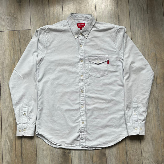 Supreme SS19 Oxford Shirt is a light blue button-up shirt from the Spring/Summer 2019 collection, released on February 21, 2019. It features a button-front closure, a standard collar, and a single chest pocket with a small Supreme tag attached to the side.