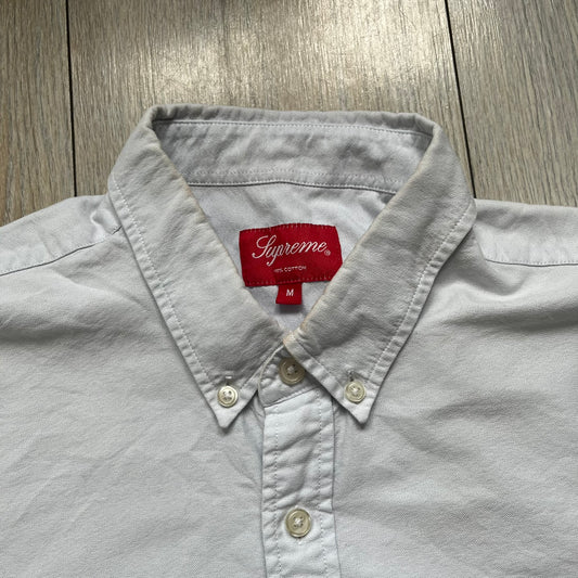 Supreme SS19 Oxford Shirt is a light blue button-up shirt from the Spring/Summer 2019 collection, released on February 21, 2019. It features a button-front closure, a standard collar, and a single chest pocket with a small Supreme tag attached to the side.