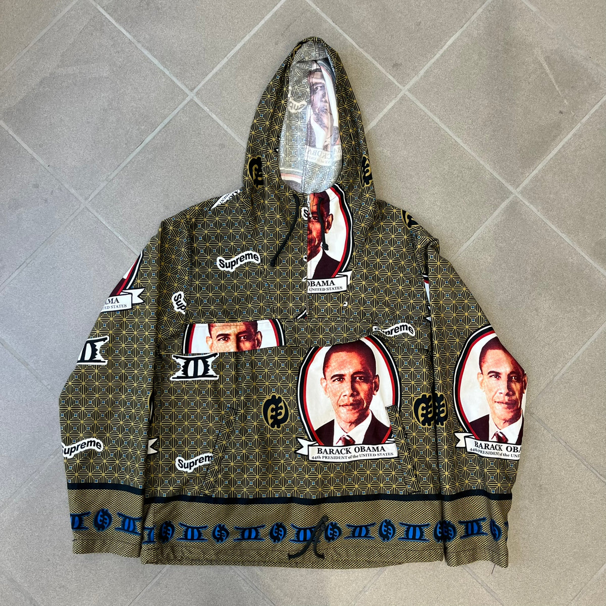 Supreme SS2017 Obama Green Anorak is a green/olive 100% cotton quarter-zip anorak with an all-over patchwork print incorporating Barack Obama’s face, the words “Barack Obama,” and scattered Supreme logos. The design pays homage to the “khanas,” vibrant patterned garments with political graphics made by local designers in Ghana to commemorate the former president’s 2009 visit. It has a drawstring hood and hem and lower front hand pockets.