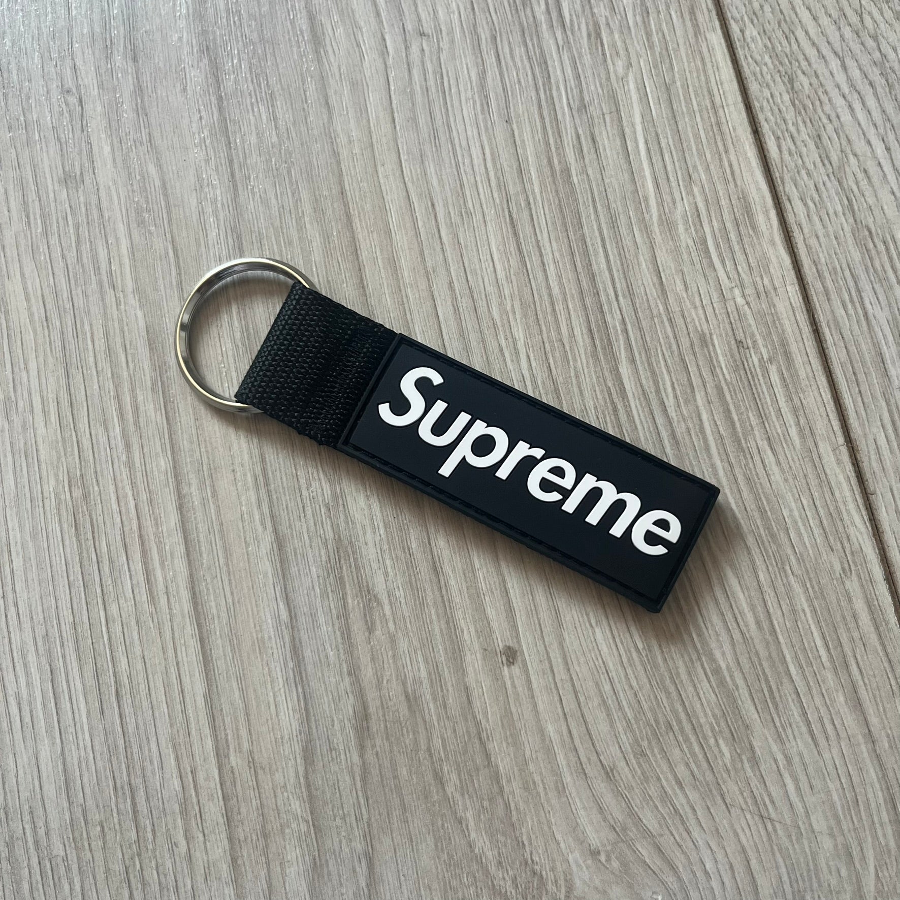 Supreme SS26 Webbing Keychain is a black keychain from the Spring/Summer 2026 collection. It features a silver-tone 1-inch keyring and a black rubber Supreme box logo patch attached to the front. The reverse side is plain with no additional graphics.