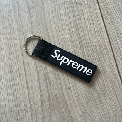 Supreme SS26 Webbing Keychain is a black keychain from the Spring/Summer 2026 collection. It features a silver-tone 1-inch keyring and a black rubber Supreme box logo patch attached to the front. The reverse side is plain with no additional graphics.