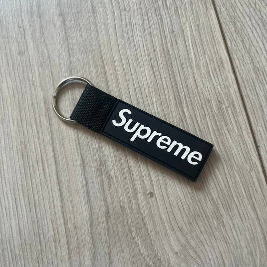 Supreme SS26 Webbing Keychain is a black keychain from the Spring/Summer 2026 collection. It features a silver-tone 1-inch keyring and a black rubber Supreme box logo patch attached to the front. The reverse side is plain with no additional graphics.