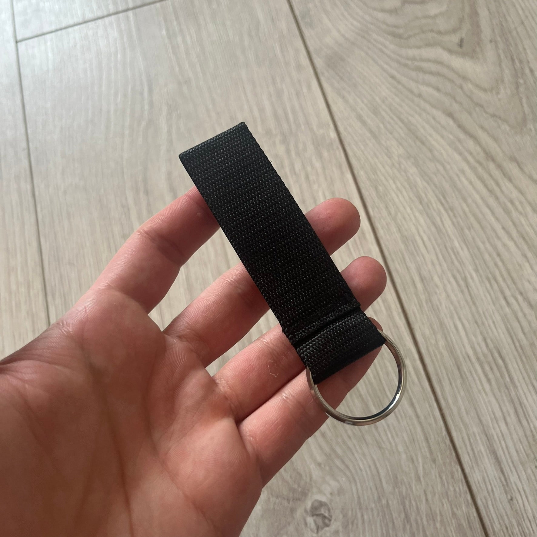 Supreme SS26 Webbing Keychain is a black keychain from the Spring/Summer 2026 collection. It features a silver-tone 1-inch keyring and a black rubber Supreme box logo patch attached to the front. The reverse side is plain with no additional graphics.