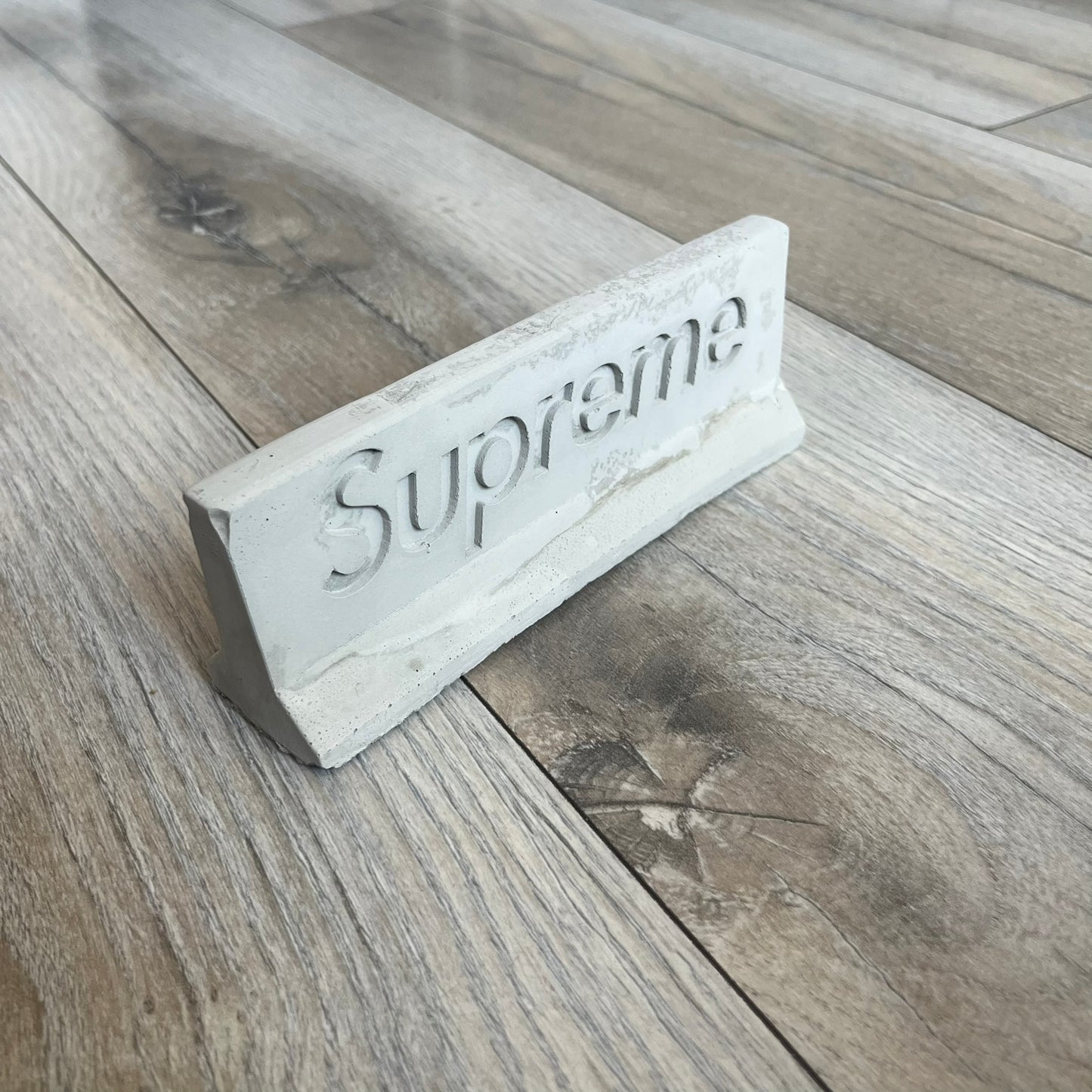 Supreme SS26 Mini Materials 1:12 Scale Jersey Barrier is a grey miniature accessory released on March 26, 2026, as part of the Spring/Summer 2026 collection. The piece replicates a standard jersey barrier in 1:12 scale with realistic construction-style detailing. This item was produced as part of Supreme’s Mini Materials accessory line.