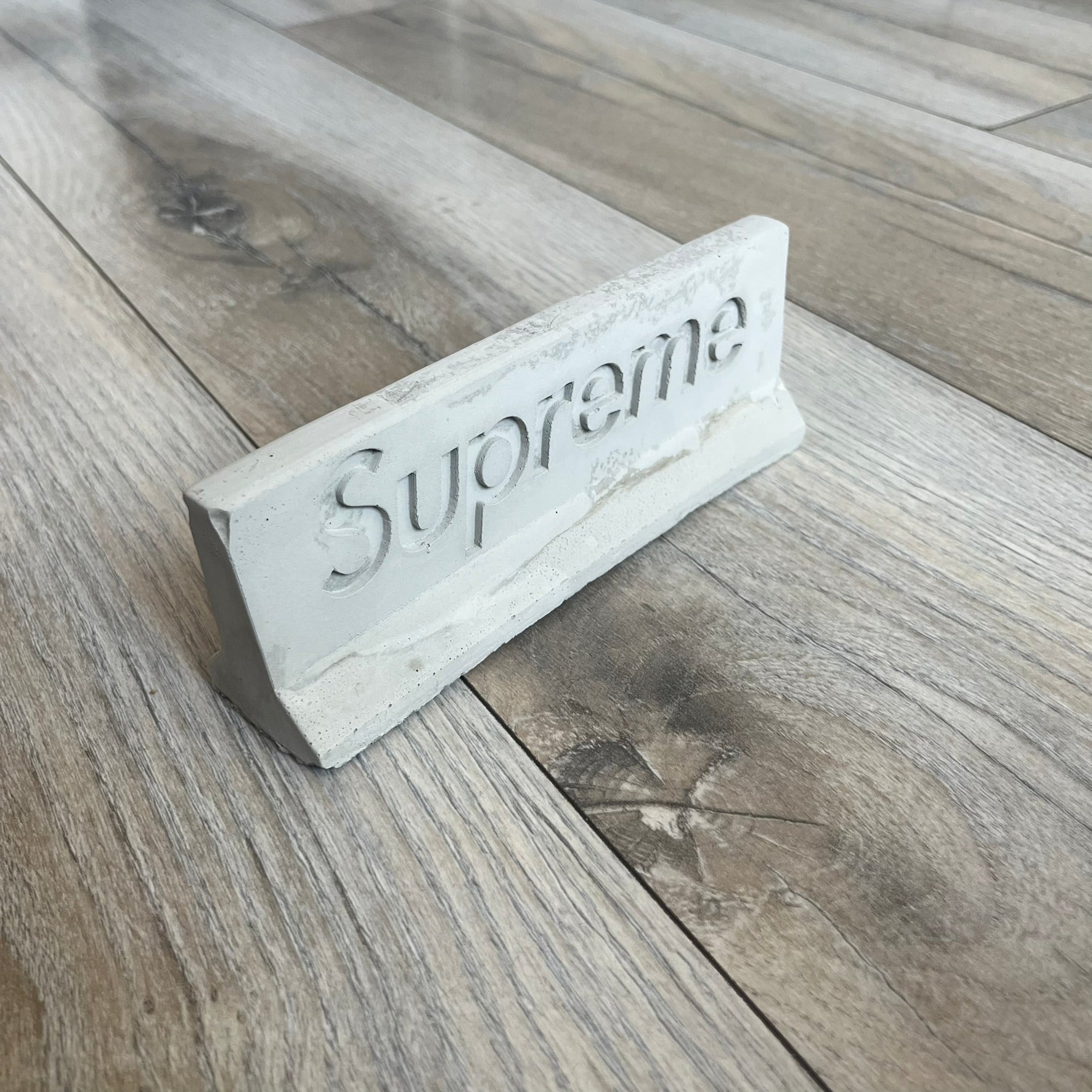 Supreme SS26 Mini Materials 1:12 Scale Jersey Barrier is a grey miniature accessory released on March 26, 2026, as part of the Spring/Summer 2026 collection. The piece replicates a standard jersey barrier in 1:12 scale with realistic construction-style detailing. This item was produced as part of Supreme’s Mini Materials accessory line.