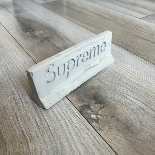 Supreme SS26 Mini Materials 1:12 Scale Jersey Barrier is a grey miniature accessory released on March 26, 2026, as part of the Spring/Summer 2026 collection. The piece replicates a standard jersey barrier in 1:12 scale with realistic construction-style detailing. This item was produced as part of Supreme’s Mini Materials accessory line.