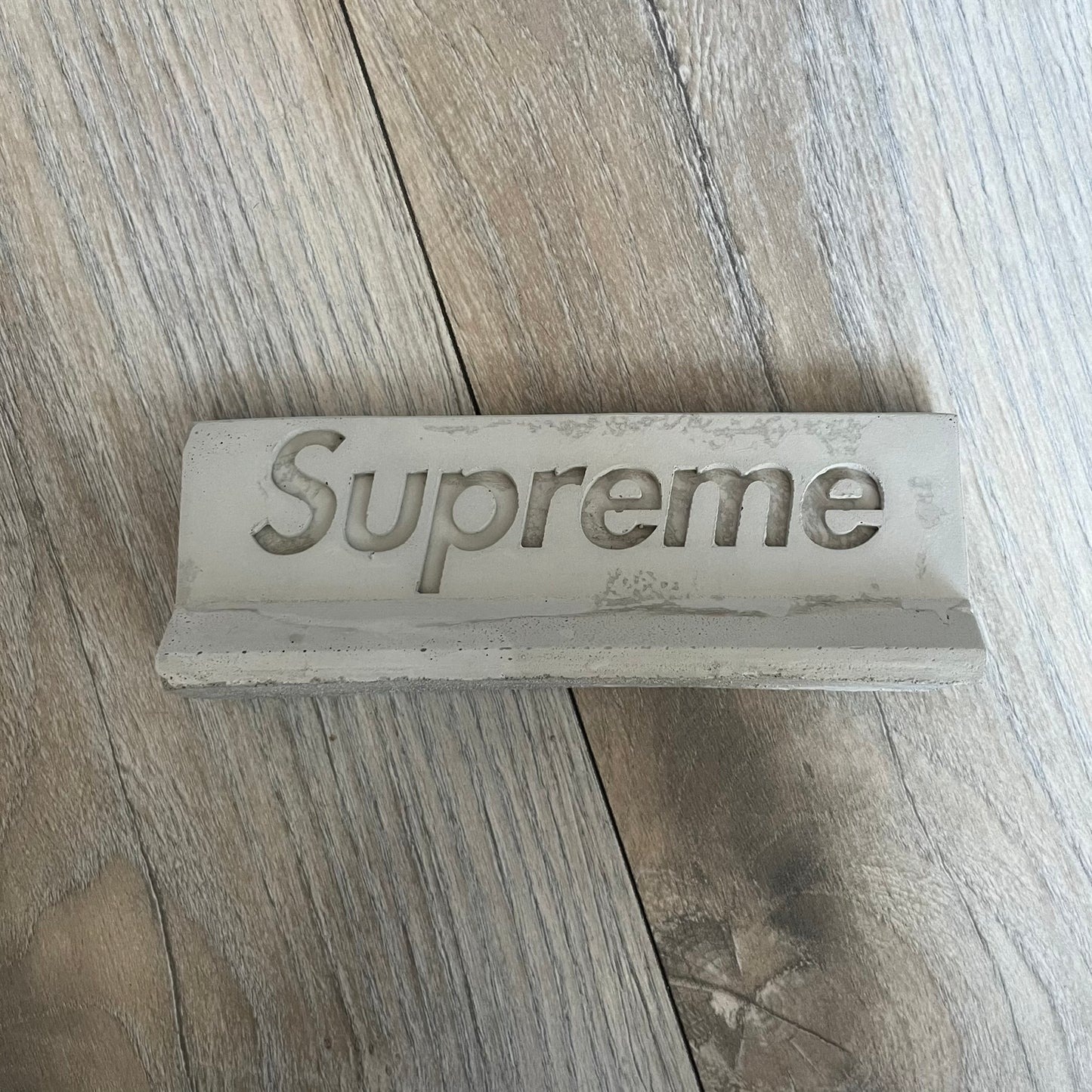 Supreme SS26 Mini Materials 1:12 Scale Jersey Barrier is a grey miniature accessory released on March 26, 2026, as part of the Spring/Summer 2026 collection. The piece replicates a standard jersey barrier in 1:12 scale with realistic construction-style detailing. This item was produced as part of Supreme’s Mini Materials accessory line.