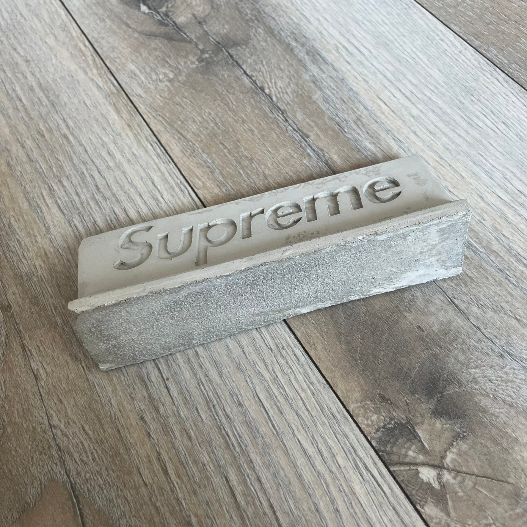 Supreme SS26 Mini Materials 1:12 Scale Jersey Barrier is a grey miniature accessory released on March 26, 2026, as part of the Spring/Summer 2026 collection. The piece replicates a standard jersey barrier in 1:12 scale with realistic construction-style detailing. This item was produced as part of Supreme’s Mini Materials accessory line.