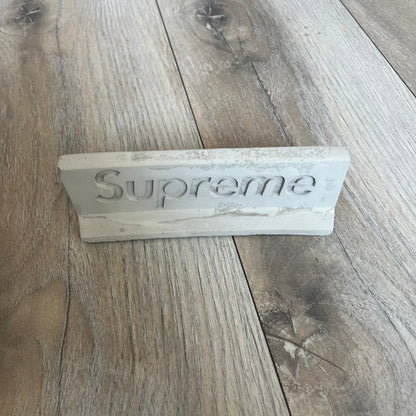 Supreme SS26 Mini Materials 1:12 Scale Jersey Barrier is a grey miniature accessory released on March 26, 2026, as part of the Spring/Summer 2026 collection. The piece replicates a standard jersey barrier in 1:12 scale with realistic construction-style detailing. This item was produced as part of Supreme’s Mini Materials accessory line.