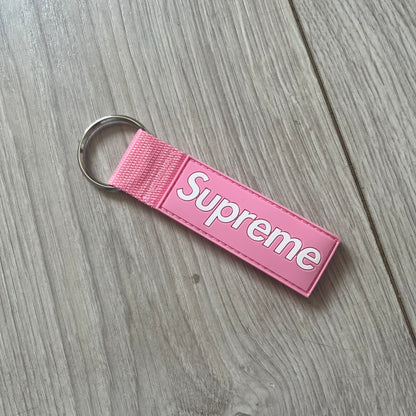  Supreme SS26 Webbing Keychain is a pink keychain from the Spring/Summer 2026 collection. It features a silver-tone 1-inch keyring and a pink rubber Supreme box logo patch attached to the front. The reverse side is plain with no additional graphics.