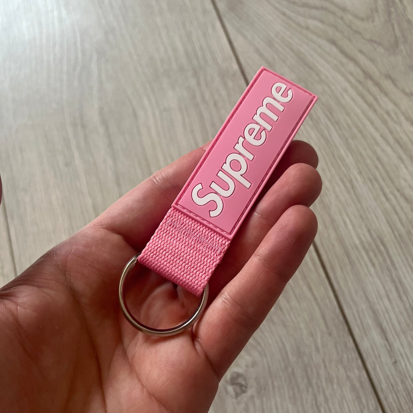  Supreme SS26 Webbing Keychain is a pink keychain from the Spring/Summer 2026 collection. It features a silver-tone 1-inch keyring and a pink rubber Supreme box logo patch attached to the front. The reverse side is plain with no additional graphics.