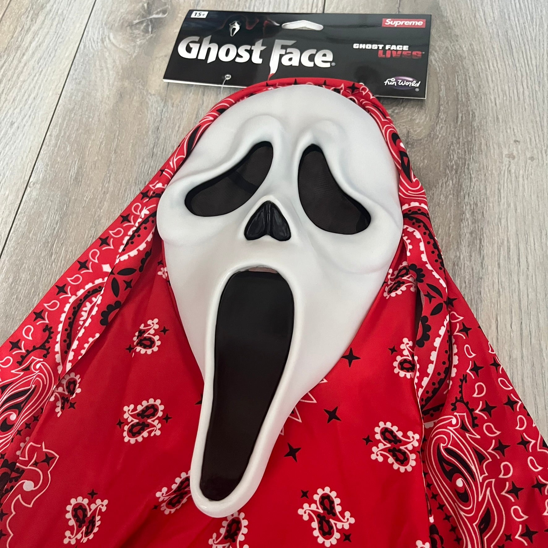 Supreme SS26 Ghostface Mask is a PVC face mask released on April 30, 2026, as part of the Spring/Summer 2026 collection. The mask features the classic white Ghostface design, combined with a red fabric hood element covered in white and black bandana-style patterns and integrated Supreme tags. Additional hanging strips on the sides enhance the hooded visual effect.