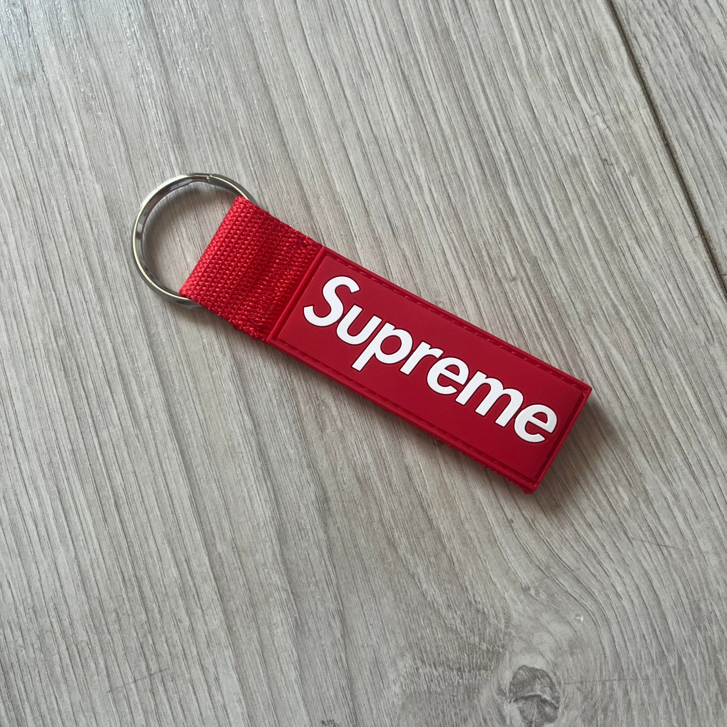 Supreme SS26 Webbing Keychain is a red keychain from the Spring/Summer 2026 collection. It features a silver-tone 1-inch keyring and a red rubber Supreme box logo patch attached to the front. The reverse side is plain with no additional graphics.