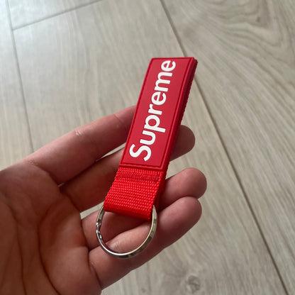 Supreme SS26 Webbing Keychain is a red keychain from the Spring/Summer 2026 collection. It features a silver-tone 1-inch keyring and a red rubber Supreme box logo patch attached to the front. The reverse side is plain with no additional graphics.
