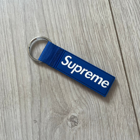 Supreme SS26 Webbing Keychain is a blue keychain from the Spring/Summer 2026 collection. It features a silver-tone 1-inch keyring and a blue rubber Supreme box logo patch attached to the front. The reverse side is plain with no additional graphics.