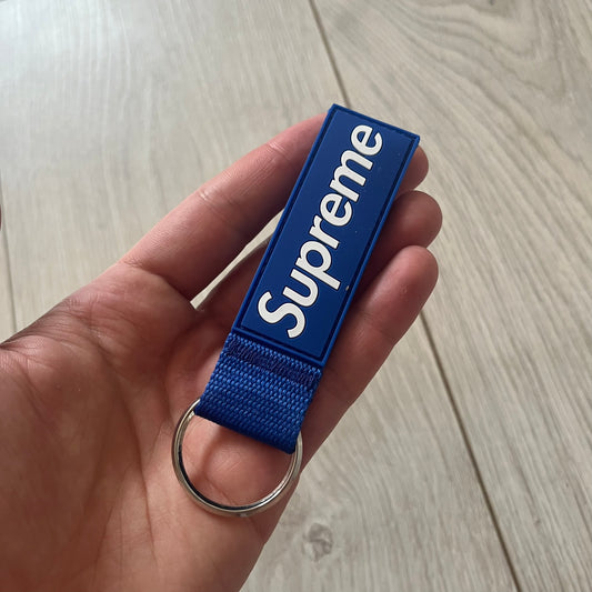 Supreme SS26 Webbing Keychain is a blue keychain from the Spring/Summer 2026 collection. It features a silver-tone 1-inch keyring and a blue rubber Supreme box logo patch attached to the front. The reverse side is plain with no additional graphics.