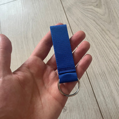 Supreme SS26 Webbing Keychain is a blue keychain from the Spring/Summer 2026 collection. It features a silver-tone 1-inch keyring and a blue rubber Supreme box logo patch attached to the front. The reverse side is plain with no additional graphics.
