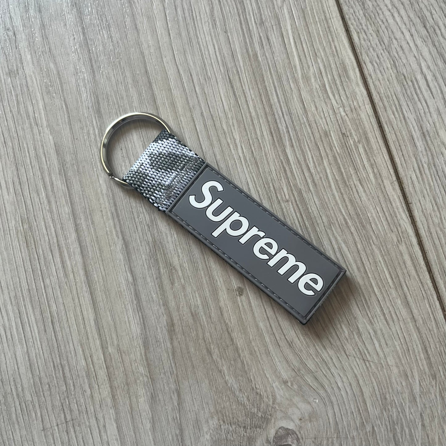 Supreme SS26 Webbing Keychain is a gray keychain from the Spring/Summer 2026 collection. It features a silver-tone 1-inch keyring and a gray rubber Supreme box logo patch attached to the front. The reverse side is a gray, white and black "snow camo".