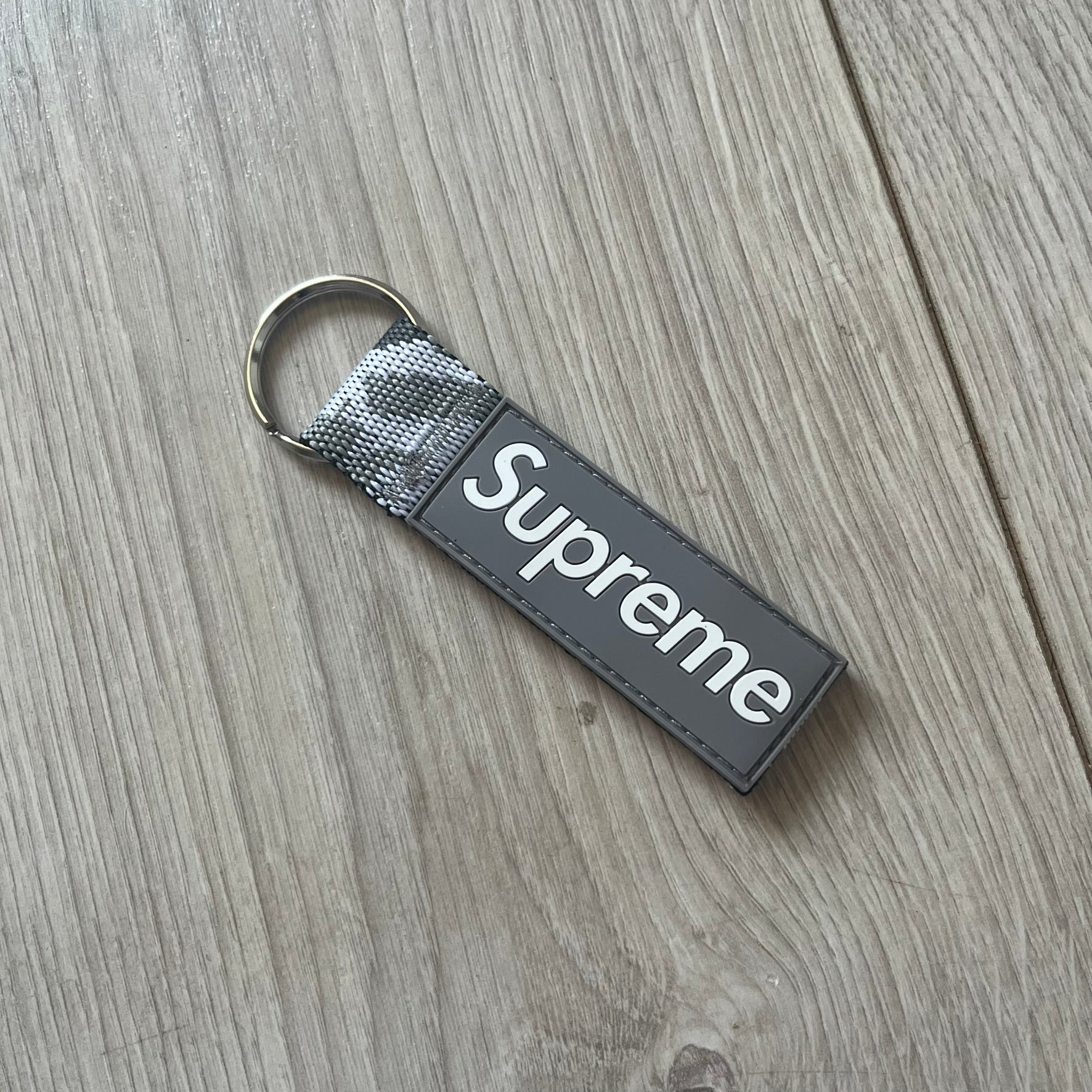 Supreme SS26 Webbing Keychain is a gray keychain from the Spring/Summer 2026 collection. It features a silver-tone 1-inch keyring and a gray rubber Supreme box logo patch attached to the front. The reverse side is a gray, white and black "snow camo".
