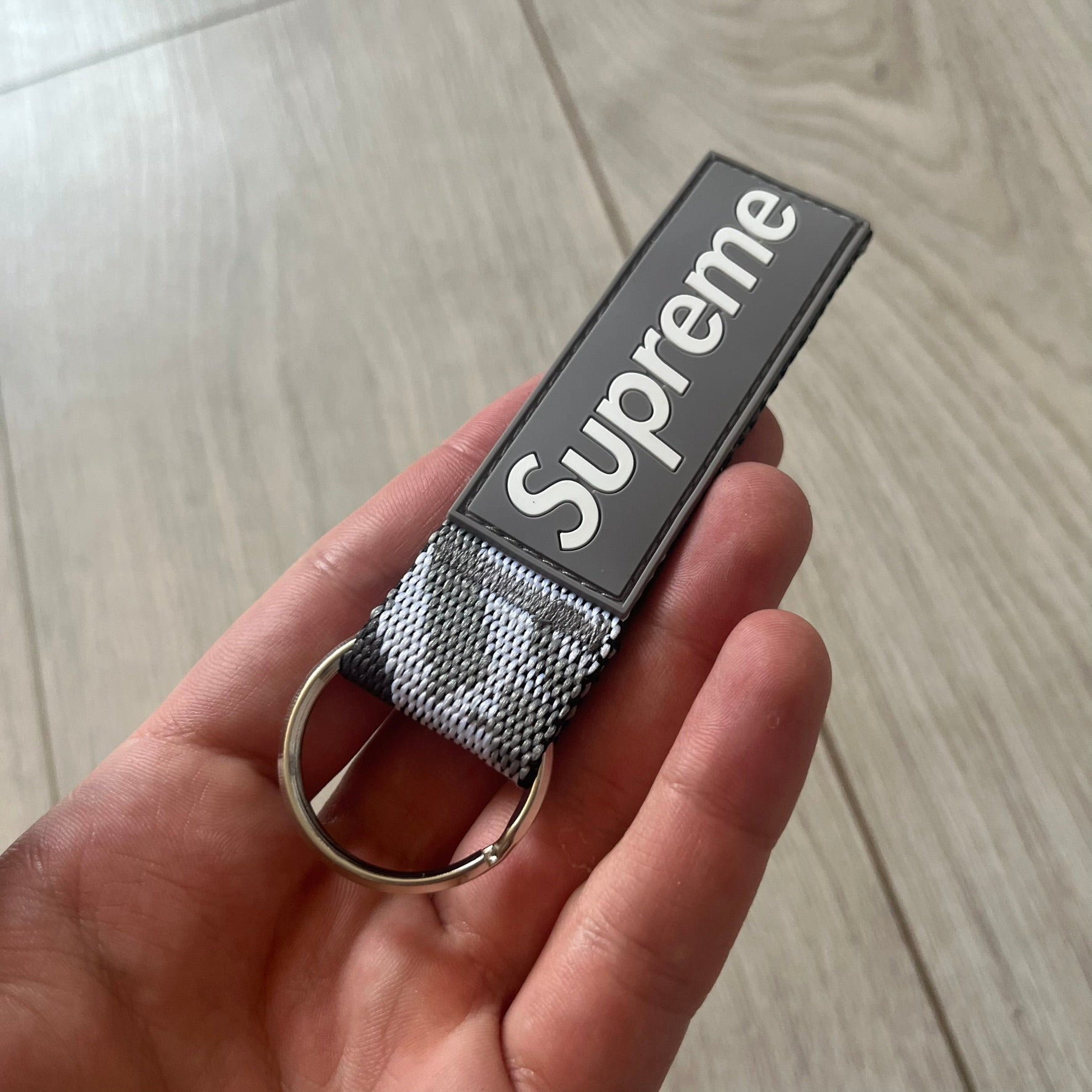 Supreme SS26 Webbing Keychain is a gray keychain from the Spring/Summer 2026 collection. It features a silver-tone 1-inch keyring and a gray rubber Supreme box logo patch attached to the front. The reverse side is a gray, white and black "snow camo".